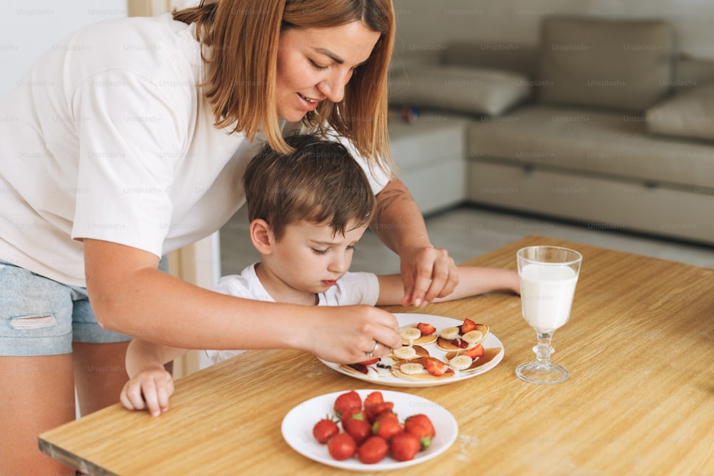 Cute toddler boy with his mother having breakfast with puncakes and berries and glass of milk in kitchen at home