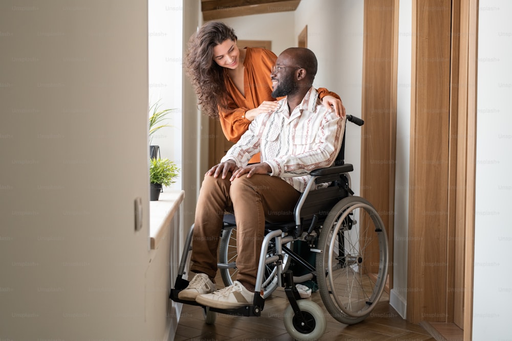 Happy young man with disability looking at his affectionate caregiver while talking to her in front of window in corridor