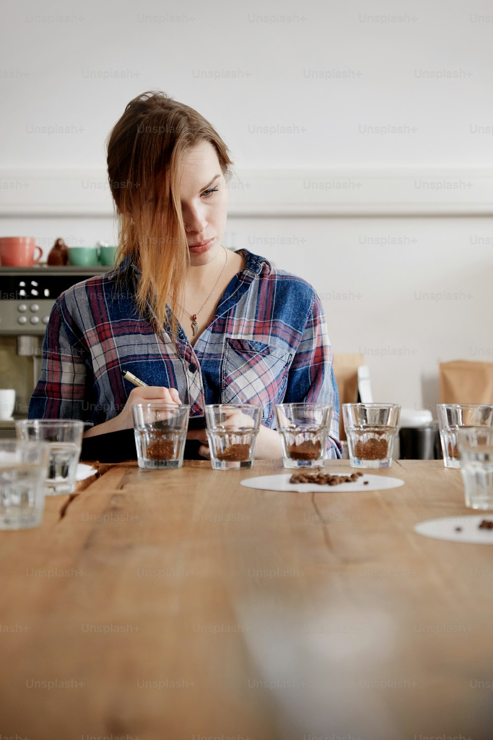 Portrait of young female barista writing result of coffee cupping test, examining cups with beans on wooden table