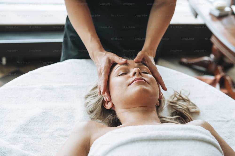 Attractive blonde young woman on couch with white linen enjoys hand facial massage in spa, treat yourself