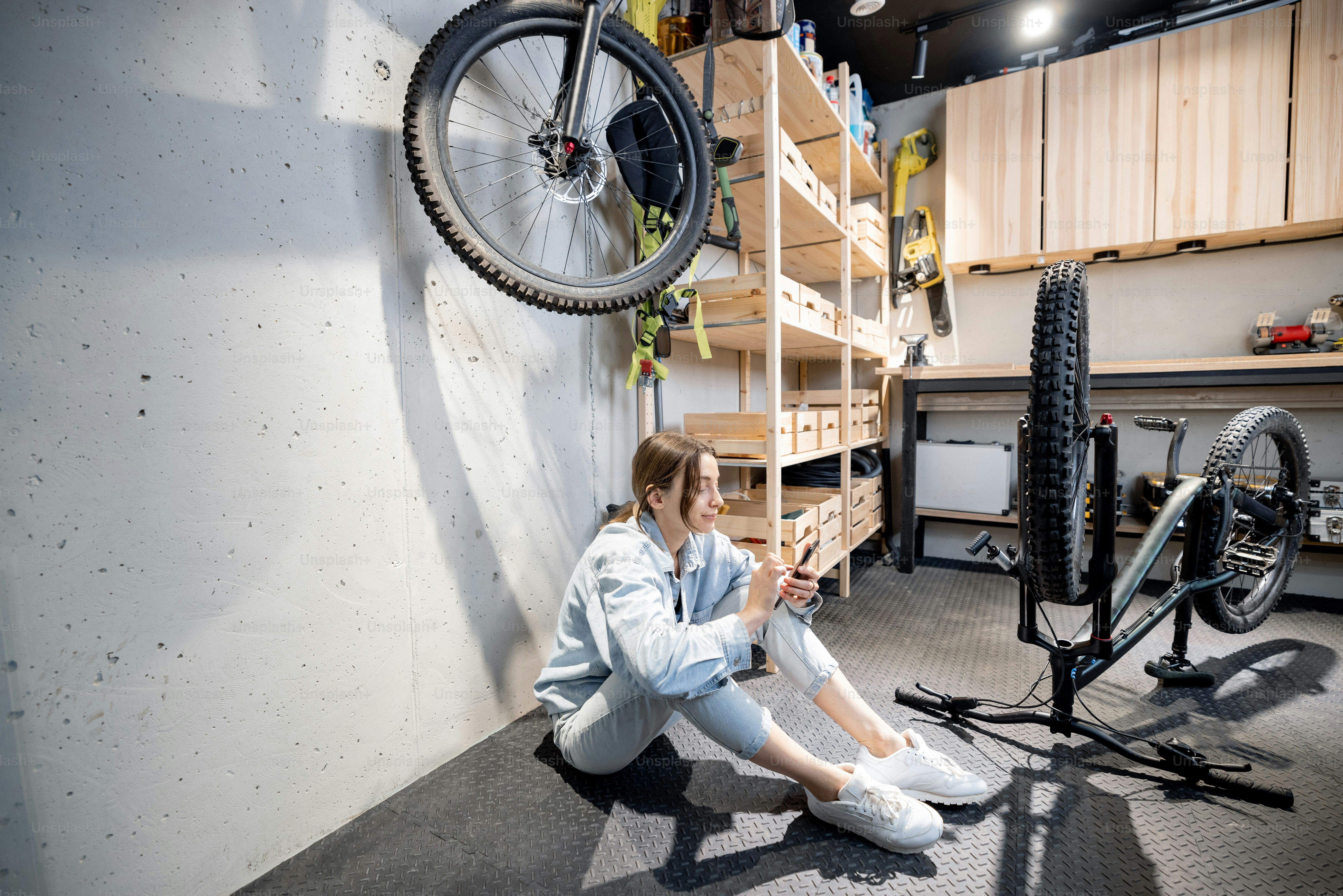 Young relaxed handywoman sitting with phone while repairing her bicycle in the well equipped workshop at home. DIY concept