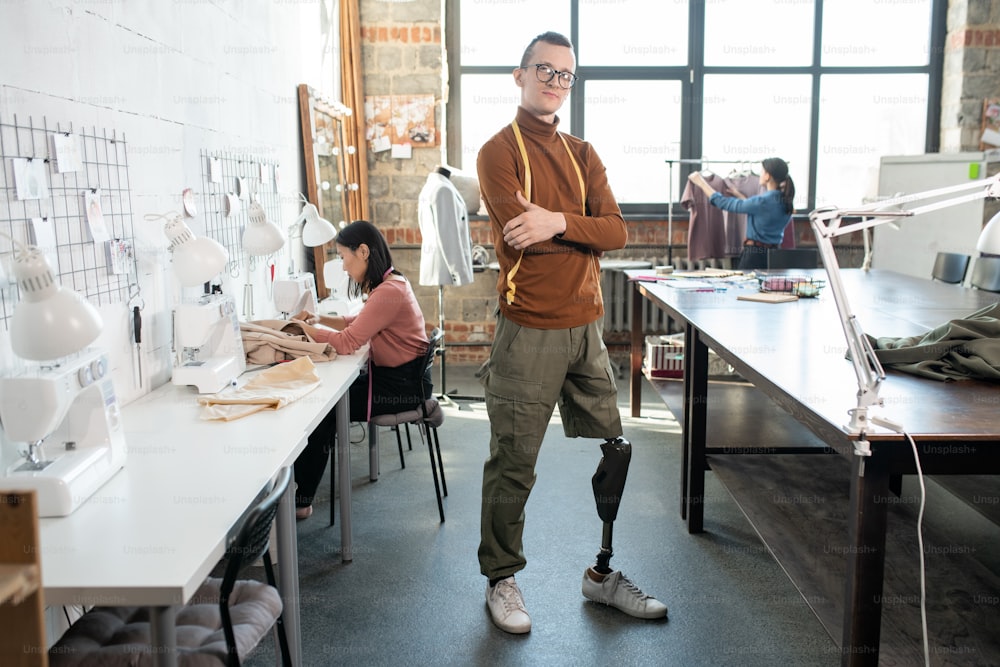 Young successful male fashion designer or tailor with prosthetic leg standing by workplace against female colleagues in large workshop