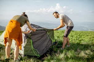 Jeune couple installant la tente sur la prairie verte, voyageant haut dans les montagnes pendant le coucher du soleil