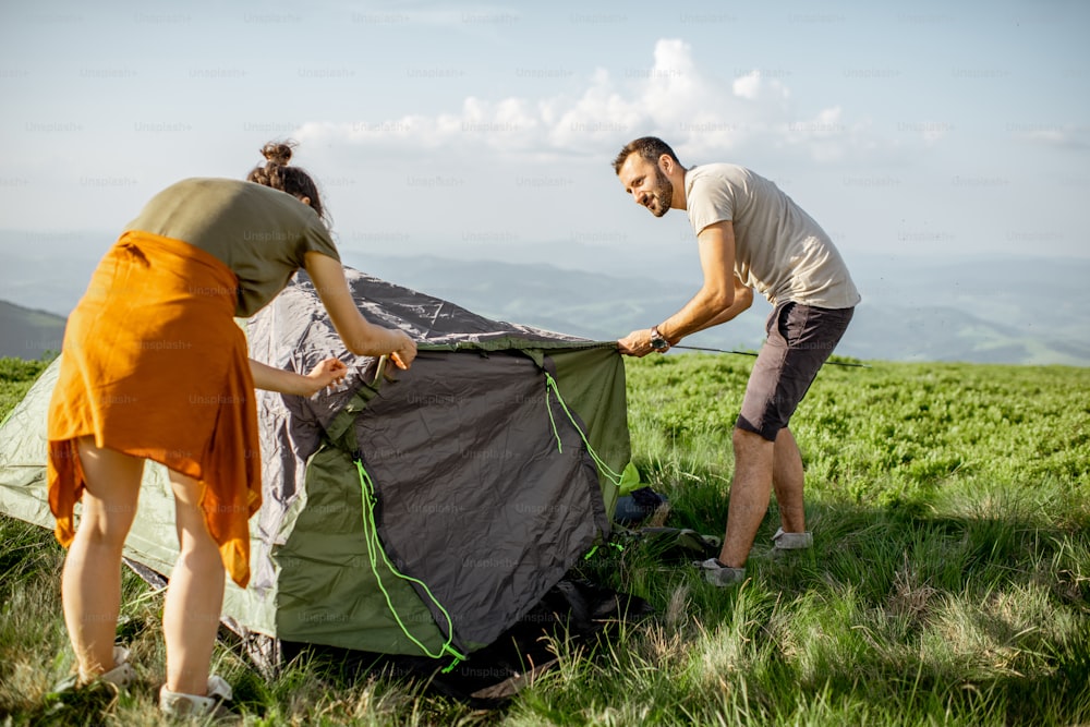 Young couple setting up the tent on the green meadow, traveling high in the mountains during the sunset