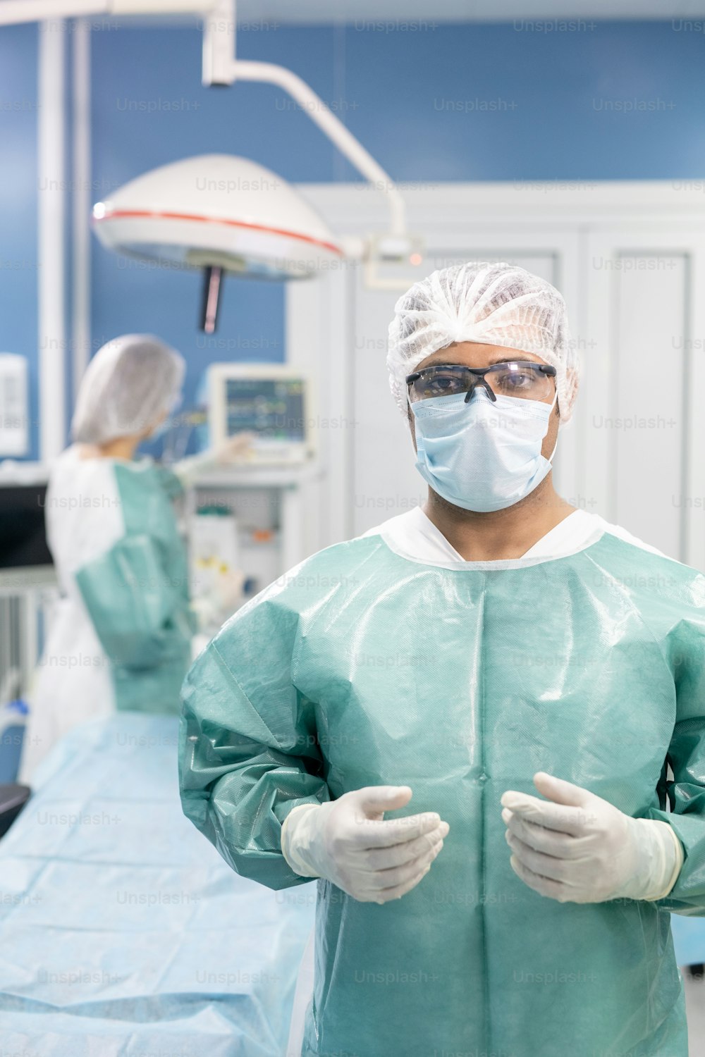 Joven cirujano profesional con overol protector, máscara, guantes y anteojos de pie en el quirófano contra su asistente