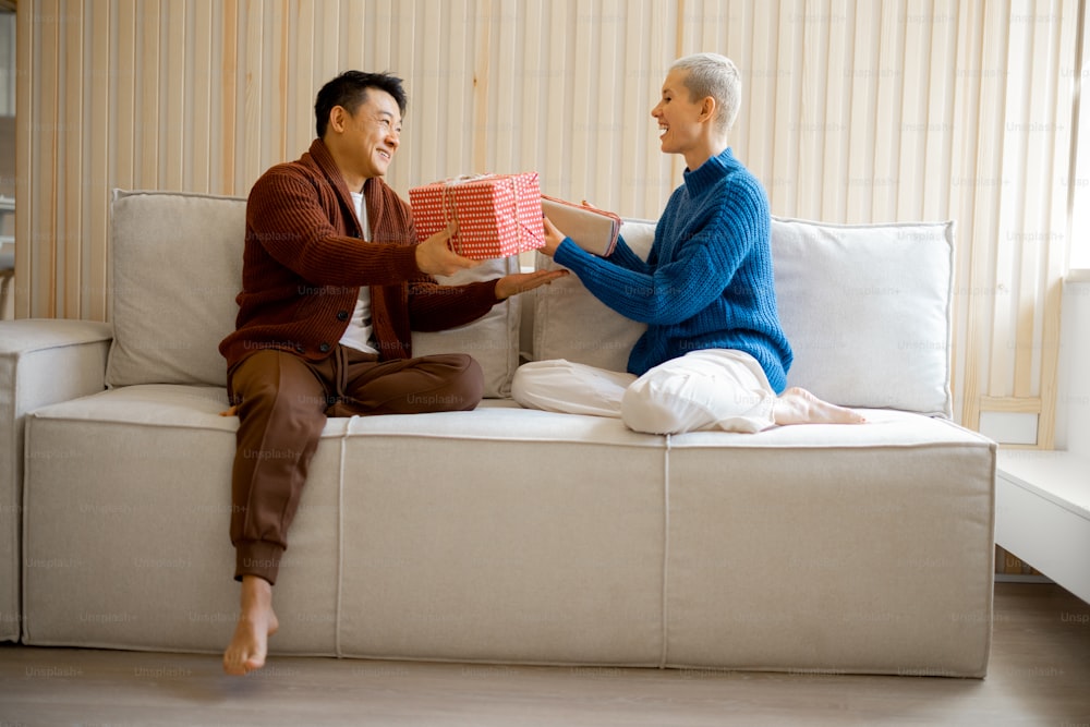 Asian man and young caucasian woman giving gifts each other. Concept of holiday, event and birthday. Idea of family relationship. Happy smiling couple sitting on sofa. Interior of modern apartment