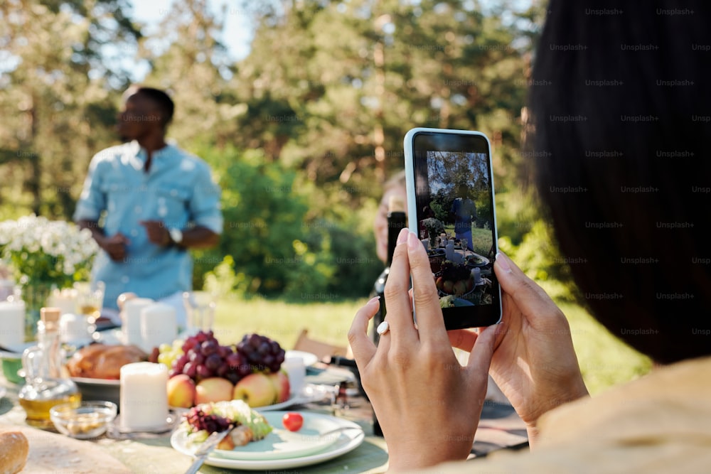 Hands of young contemporary female millennial with smartphone recording video of outdoor party with friends by served table