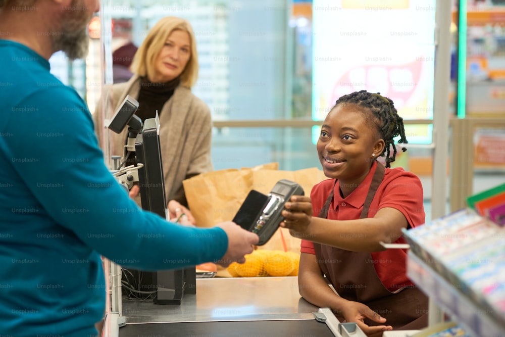 Assistente de loja feminino jovem feliz no uniforme olhando para o comprador masculino maduro em pulôver azul pagando por mercadorias por cartão de crédito