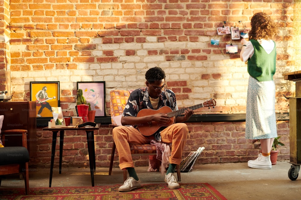 Jeune homme assis dans un fauteuil et jouant de la guitare acoustique tandis que sa petite amie debout devant un mur de briques et regardant des photos