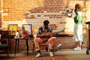 젊은 남자는 안락의자에 앉아 어쿠스틱 기타를 연주하고 그의 여자친구는 벽돌 벽 앞에 서서 사진을 보고 있다