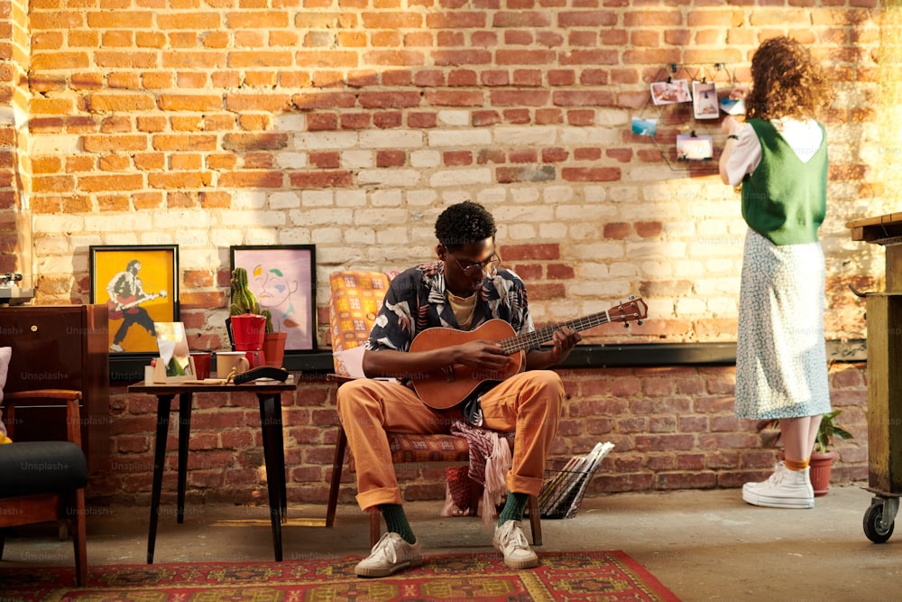 Young man sitting in armchair and playing acoustic guitar while his girlfriend standing in front of brick wall and looking at photos