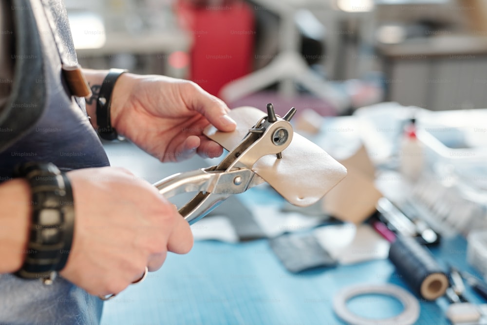 Hands of craftsman in apron with special handtool making holes in small piece of beige leather while standing by workplace