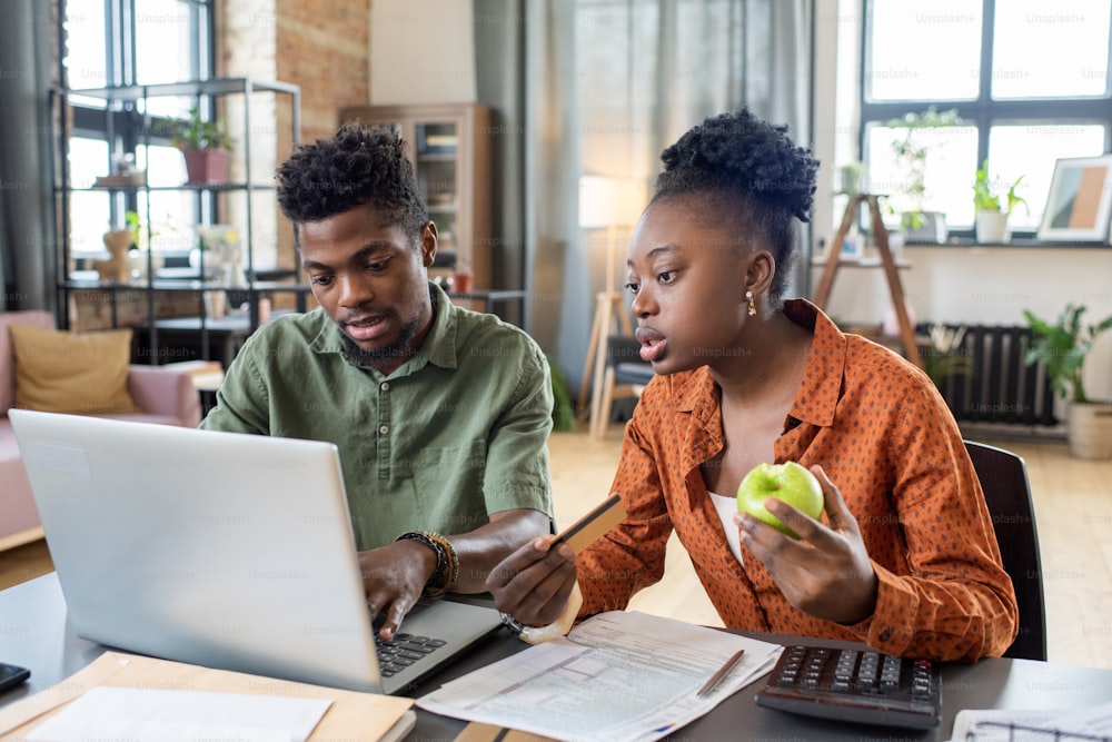 Junges afroamerikanisches Paar sitzt mit Papieren am Tisch und benutzt Laptop, während es das Steuerrückerstattungsformular online ausfüllt