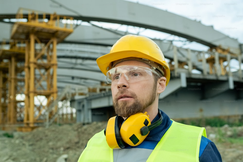 Close-up of builder in work helmet and in protective headphones standing outdoors with building under construction in the background