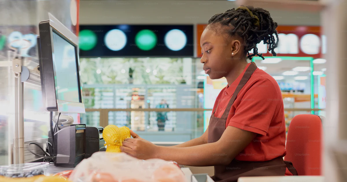 Counter Assistant Young female shop assistant in uniform scanning fresh lemons in bag ...