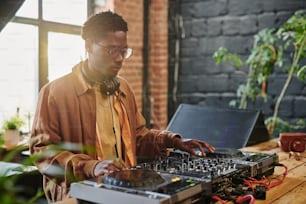 African American guy with headphones on neck touching vynil disks on musical equipment standing on table in loft apartment or studio