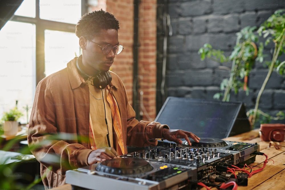 Chico afroamericano con auriculares en el cuello tocando discos de vinilo en equipos musicales de pie sobre la mesa en un loft, apartamento o estudio
