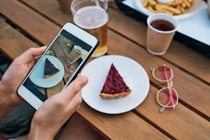 Hands of young female with smartphone taking photo of tasty cake on plate and drinks