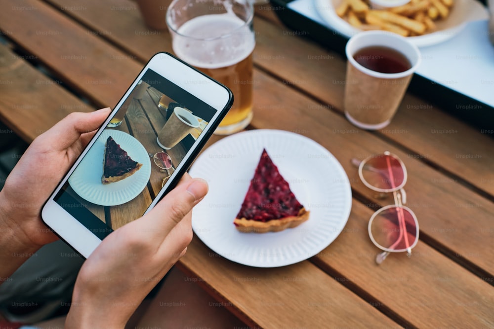 Hands of young female with smartphone taking photo of tasty cake on plate and drinks