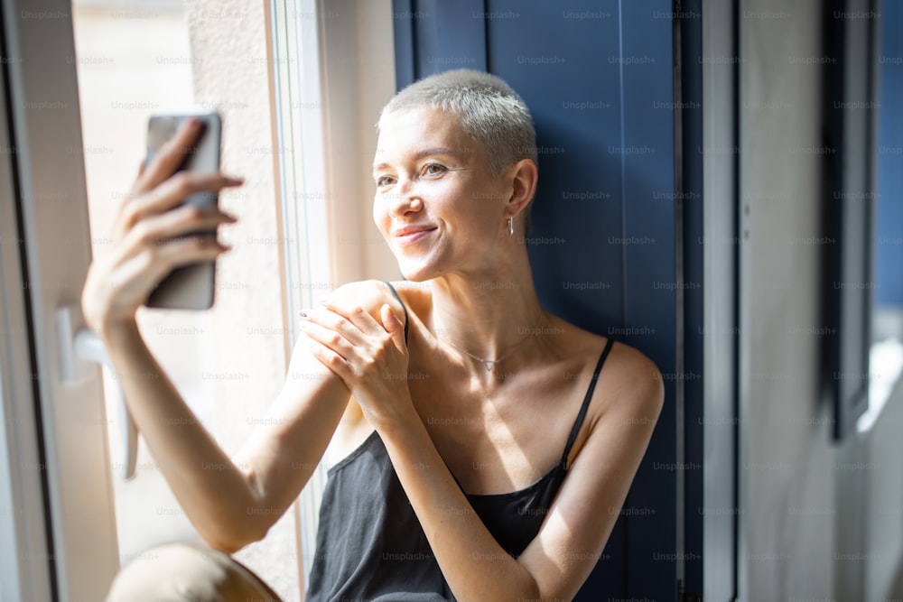La mujer feliz y elegante se divierte comunicándose por teléfono o haciendo fotos selfie mientras está sentada relajada en el alféizar de la ventana de casa.