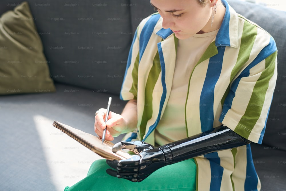 Mulher nova com mão mioelétrica segurando bloco de notas enquanto sentado no sofá na sala de estar e desenho esboço com lápis