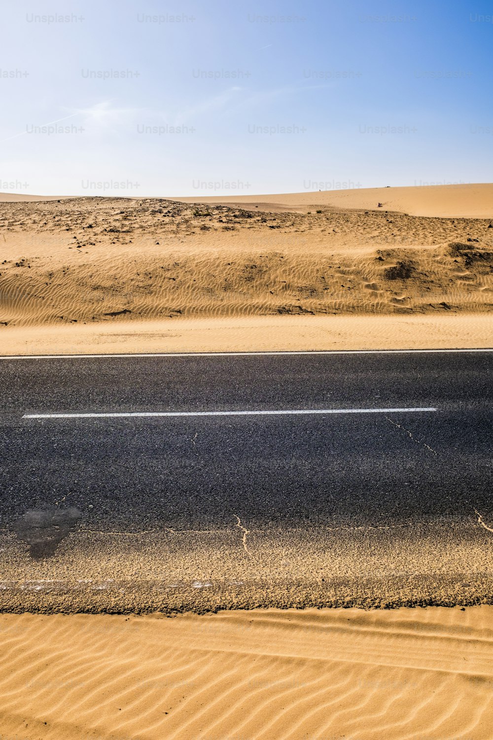 Black asphalt road with sand dunes and desert on left and right and blue sky in background  - travel concept in arid desertic world due a bad climate change future - water emergency planet