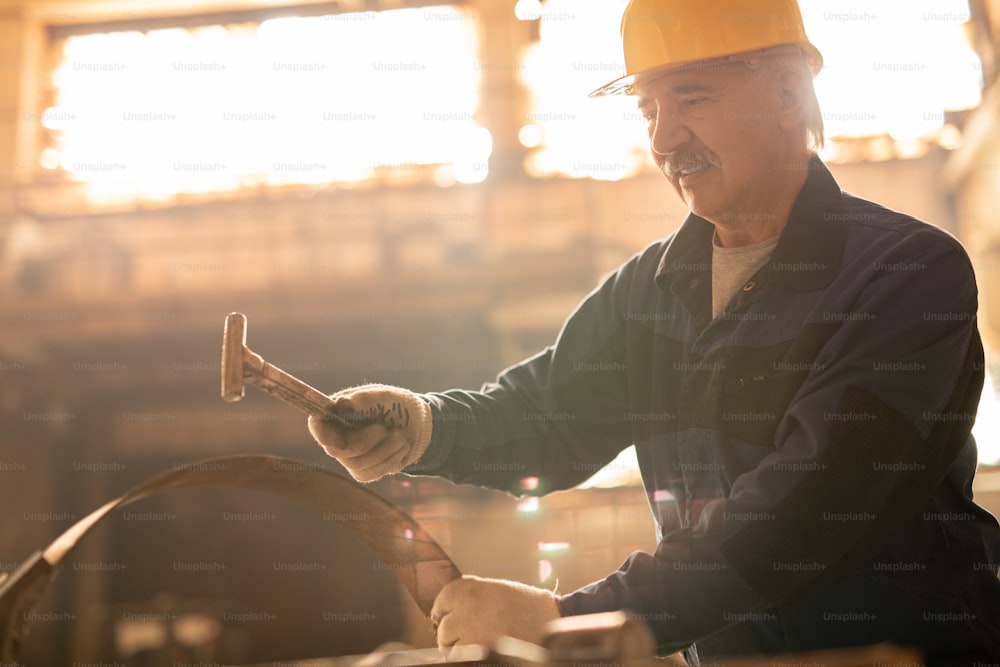 Senior engineer or machinebuilder in helmet, gloves and workwear hammering metallic detail or workpiece