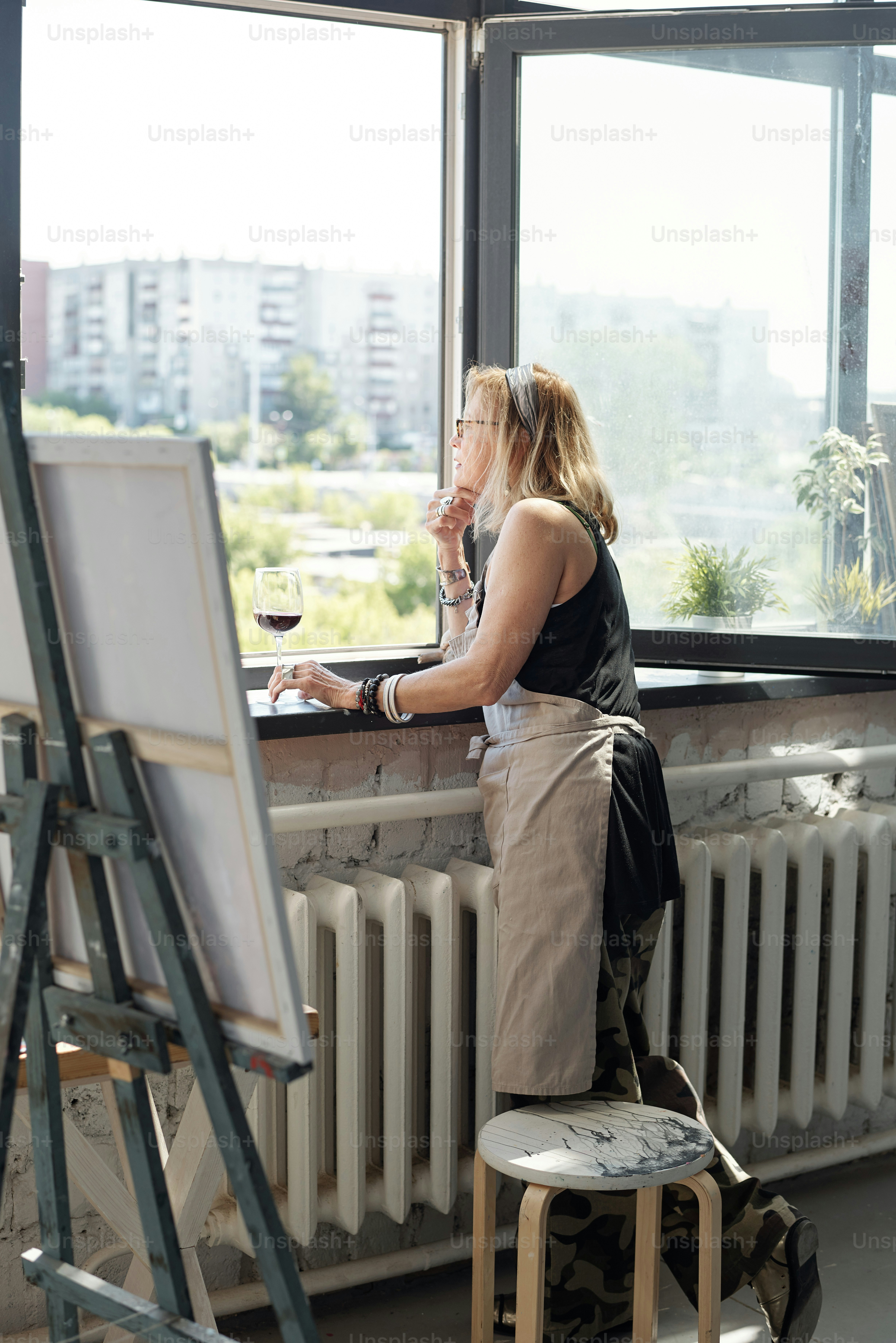 Pensive mature artist in apron contemplating cityscape through window while finding inspiration in own studio