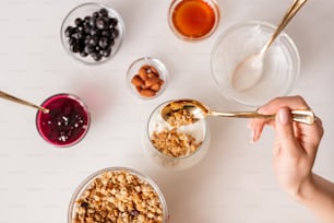Hand of young woman with teaspoon putting muesli into glass with fresh sourcream while making yoghurt with jam, almond nuts, honey and berries