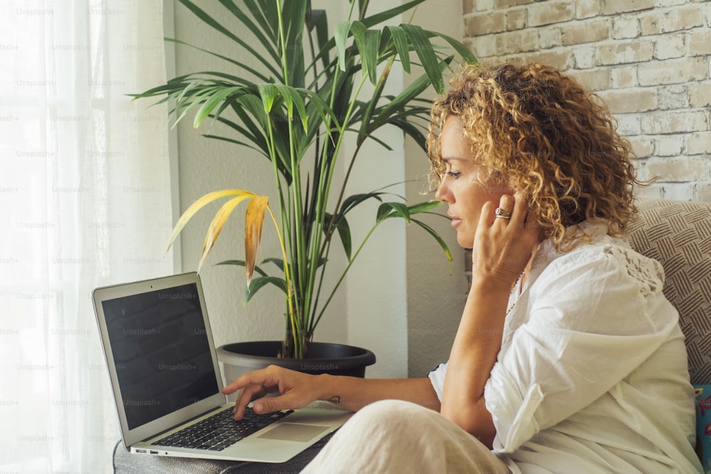 Side view of young modern adult people caucasian female using laptop sitting at home. Woman with blonde curly hair working on computer in smart job activity sitting on the sofa in indoor leisure