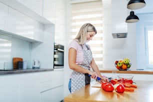 Lächelnde attraktive, würdige kaukasische blonde Frau in Schürze, die Gemüse schneidet, während sie in der Küche steht. Auf der Küchentheke stehen Karotten, Tomaten und Paprika.
