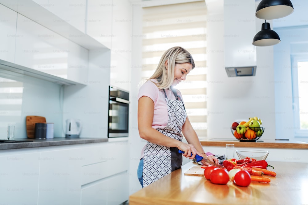 Sonriente atractiva digna mujer rubia caucásica en delantal cortando verduras mientras está de pie en la cocina. En la encimera de la cocina hay zanahorias, tomates y pimientos.