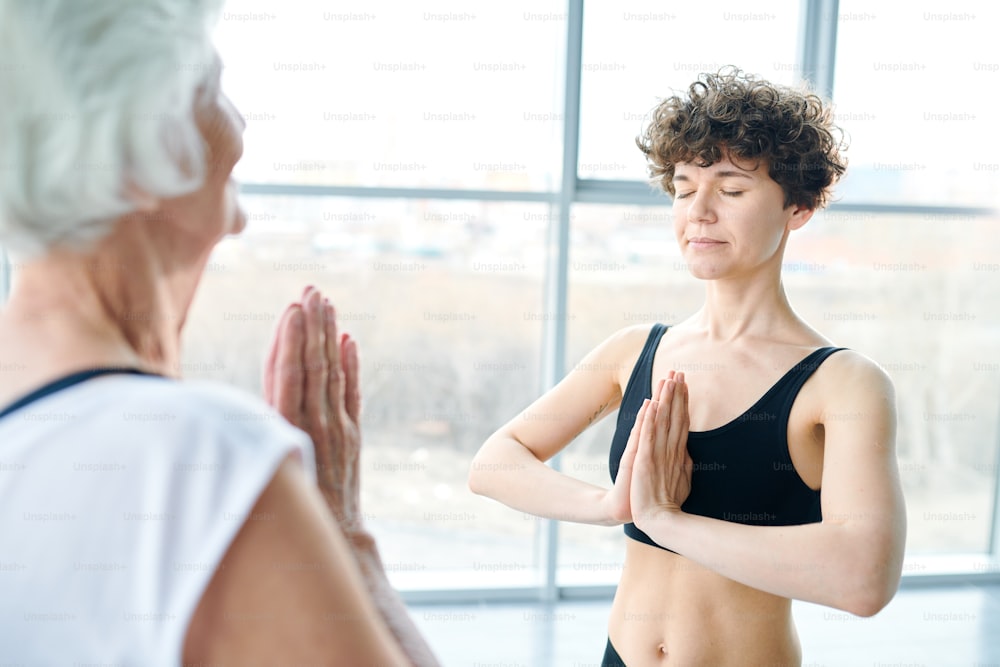 Young woman with her hands put together by chest standing in front of senior female while both practicing yoga