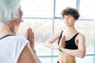 Mulher jovem com as mãos juntas pelo peito em pé na frente da fêmea sênior enquanto ambas praticam yoga