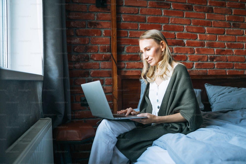 Mujer adulta de cuarenta años freelancer con cabello largo rubio en ropa casual usando computadora portátil sentada en la cama en casa