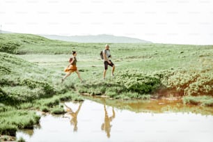 Man and woman hiking near the lake in the mountains, landscape view on the green meadow with lake during the sunny day