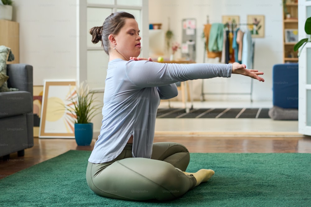 Vista lateral de una niña con síndrome de Down sentada en el suelo con las piernas cruzadas mientras practica ejercicio de yoga o posición en la sala de estar