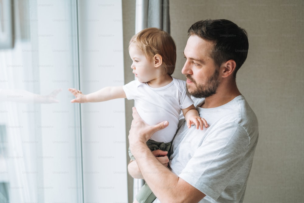 Young man father with baby girl on window sill looking at window at home