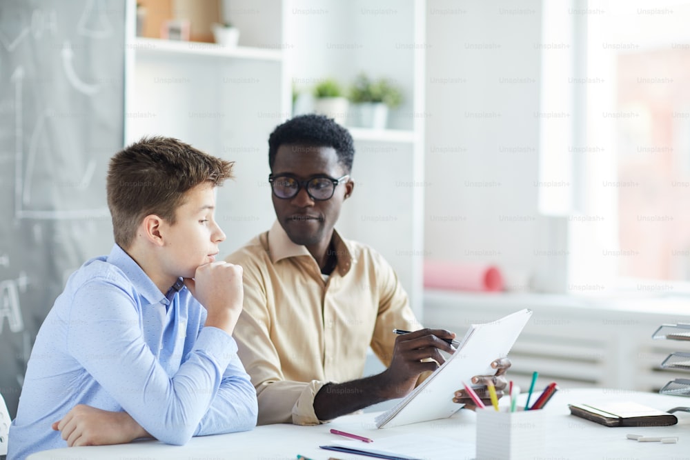 Clever schoolboy commenting his essay while teacher pointing at paper and listening to him