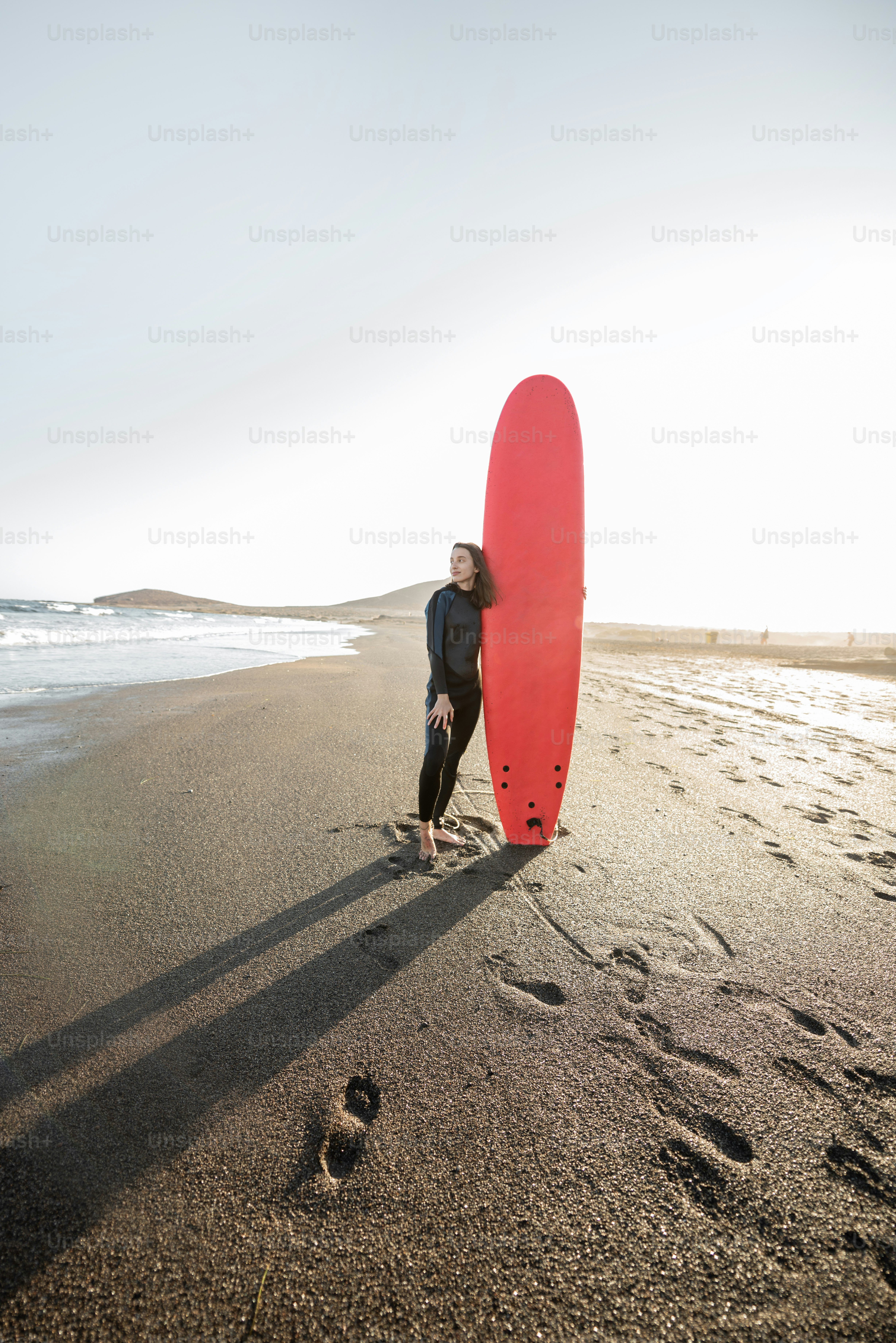 Portrait of a young woman in wetsuit standing with red surfboard on the ...