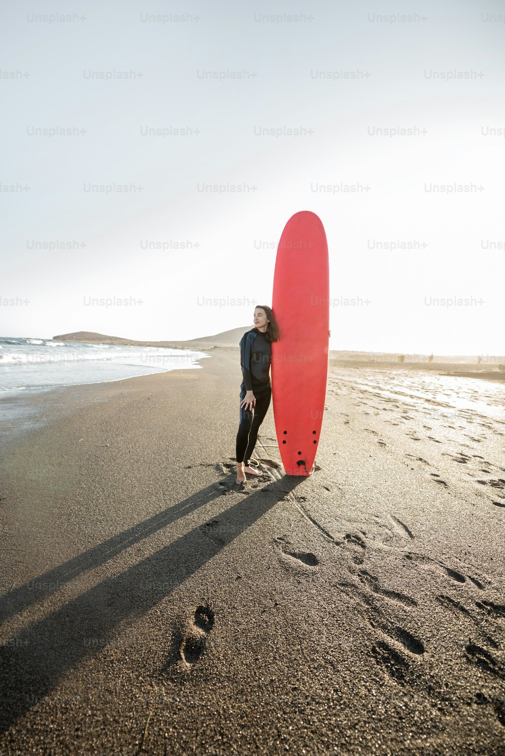 Portrait of a young woman in wetsuit standing with red surfboard on the ocean beach during a sunset. Water sport and active lifestyle concept