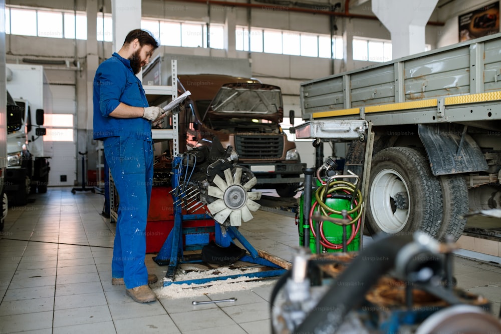 Young serious worker of automobile repair service reading instructions of transport maintenace while standing in workshop