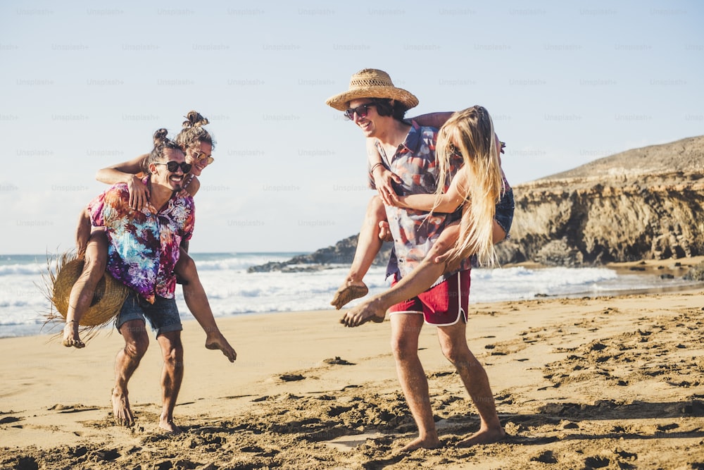 행복한 젊은이들은 함께 즐거운 시간을 보낸다 - 젊은 남녀 그룹이 해변에서 여름 휴가 휴가를 즐긴다 - 남자들은 화창한 날에 여자를 어깨에 메고 있다 - 배경의 바다 - 관광 개념
