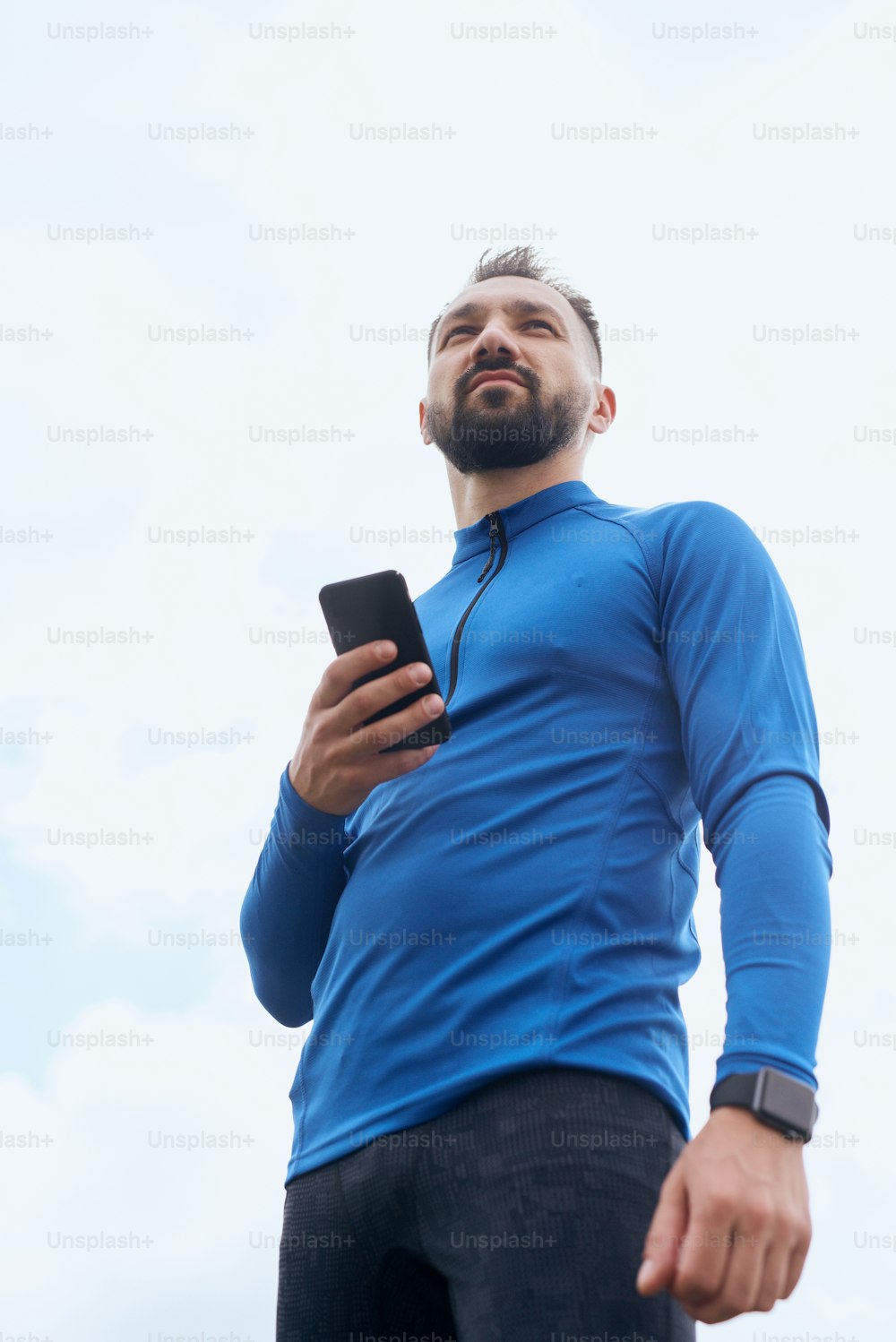 Portrait of mid age bearded sportsman checking his smart watch with smart phone, synchronizing, exercising outdoor in summer, on gloomy day with scenic view, wearing blue shirt