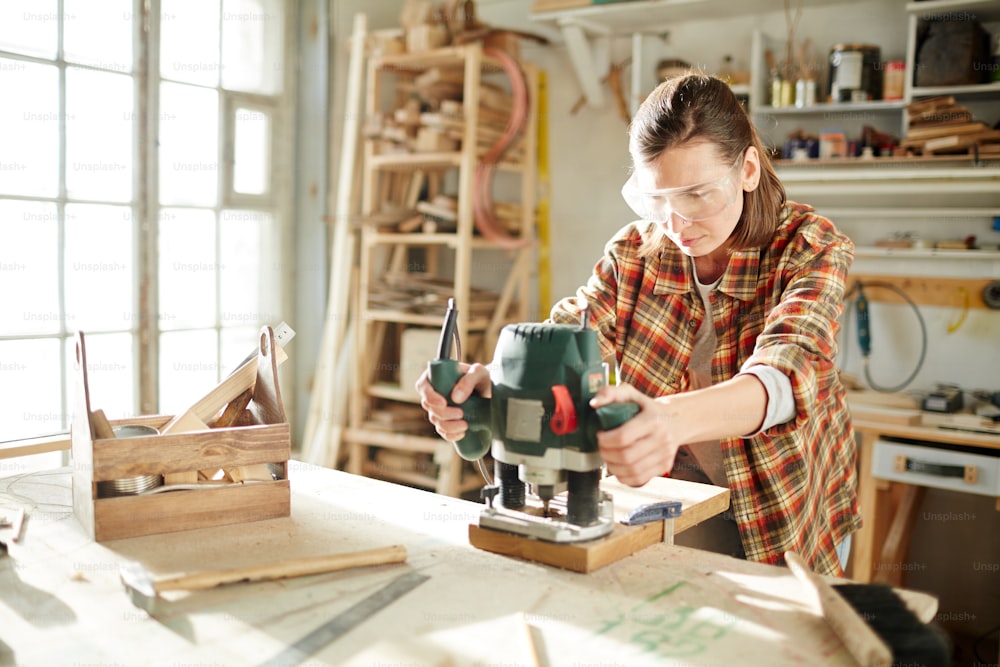 Young woman in workwear and protective eyeglasses holding by fretsaw handles while processing wooden board