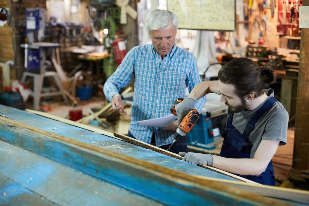 Junger Ingenieur bohrt Holzbretter, während sein älterer Kollege ihm Arbeitsanweisungen gibt