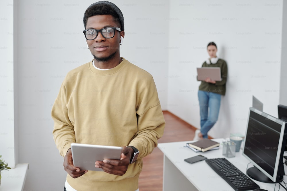 Programador masculino jovem contemporâneo com tablet olhando para a câmera contra colega de trabalho do sexo feminino com rede de laptop pela parede