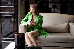 Stylish woman in green costume sits with phone on comfortable sofa at lobby bar of luxury hotel. Concept of comfortable stay on business travel