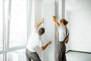 Two professional workmen in workwear installing decorative radiator in the white living room, finishing the repair in the apartment