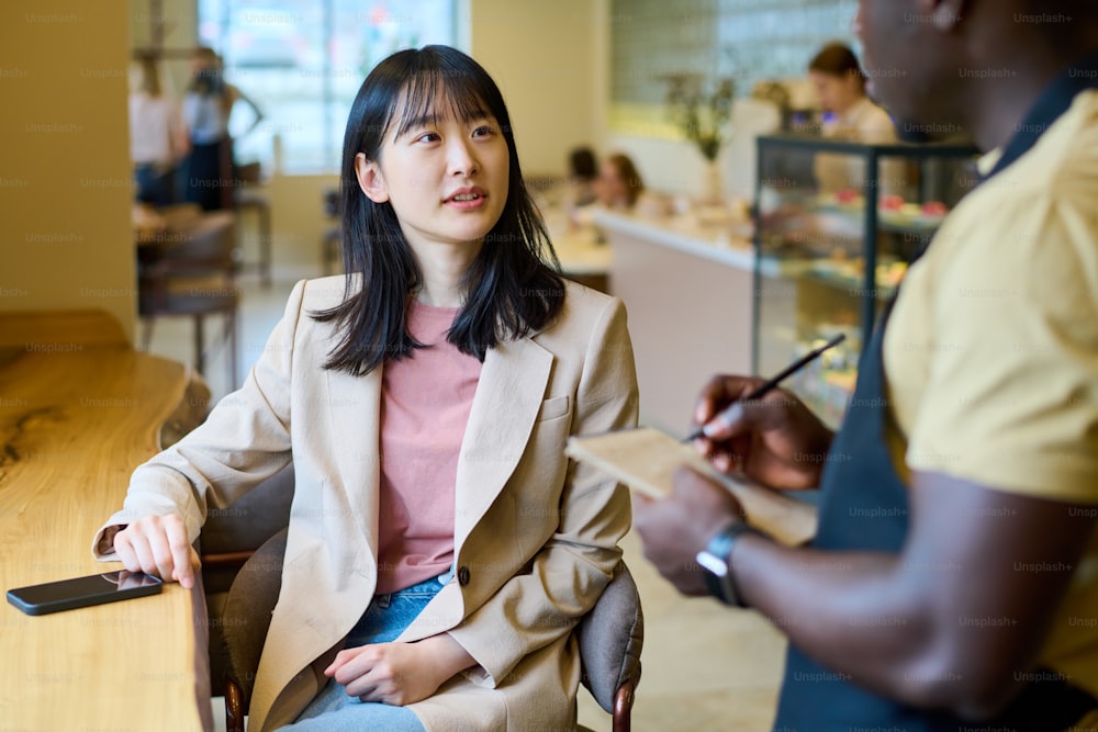 Giovane donna d'affari asiatica seduta al tavolo e parlando con il cameriere, che fa un ordine durante il pranzo in caffè
